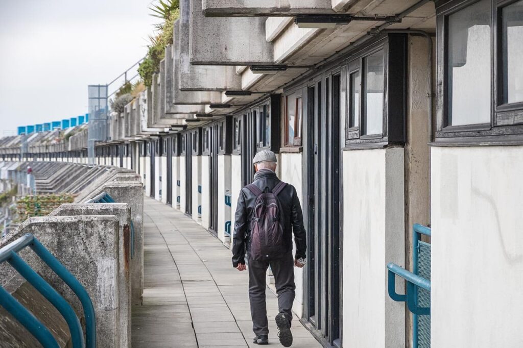 A person wearing a black jacket and carrying a backpack walks along a concrete terrace with multiple doorways and small windows. The architecture features uniform, geometric designs, and the scene is overcast. ©Mobysoft