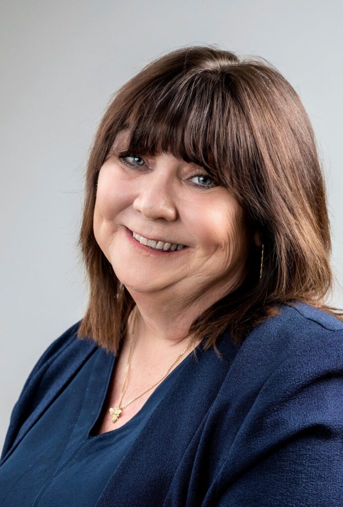 A woman with shoulder-length brown hair smiles at the camera, wearing a navy blue top and a gold necklace. The background is a light gray, creating a soft and professional look. ©Mobysoft