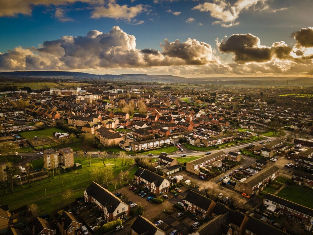 Aerial view of a suburban area during sunset, with scattered houses, green fields, and a dramatic sky filled with clouds. The landscape includes a mix of residential neighborhoods and natural scenery stretching towards distant hills. ©Mobysoft