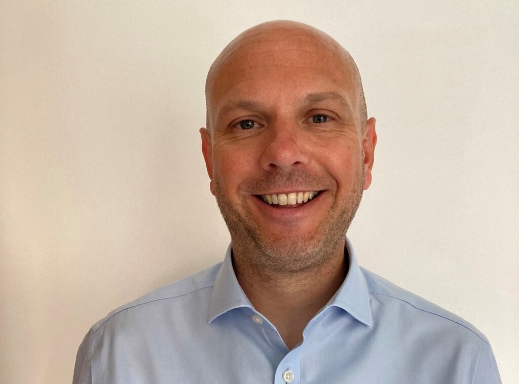 Smiling Mark Walker, a man with a shaved head and light stubble, stands confidently in a light blue button-up shirt against a plain cream background. ©Mobysoft
