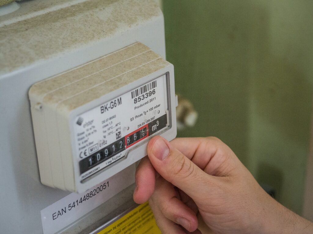 A person's hand is carefully adjusting a household gas meter with a numeric display, acutely aware of how the cost of living impacts energy consumption. The meter label shows model BK-G6M and an EAN code. ©Mobysoft