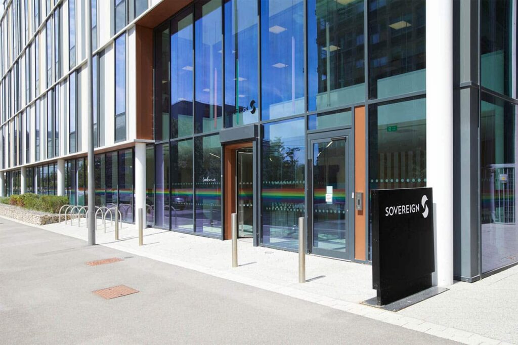 The sleek, modern glass building exterior features large windows and metal accents, embodying the spirit of intelligent automation. A sign reading "Sovereign" graces the entrance. The walkway is thoughtfully lined with plants, while bike racks enhance its functionality. ©Mobysoft