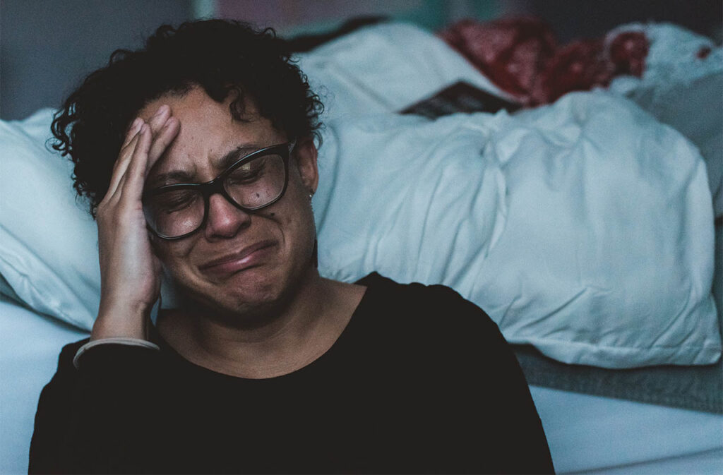 A person with glasses and curly hair sits in a dimly lit room, wearing a black sweater. They appear distressed, with their hand on their forehead and tears running down their face. A bed with a white blanket is in the background. ©Mobysoft