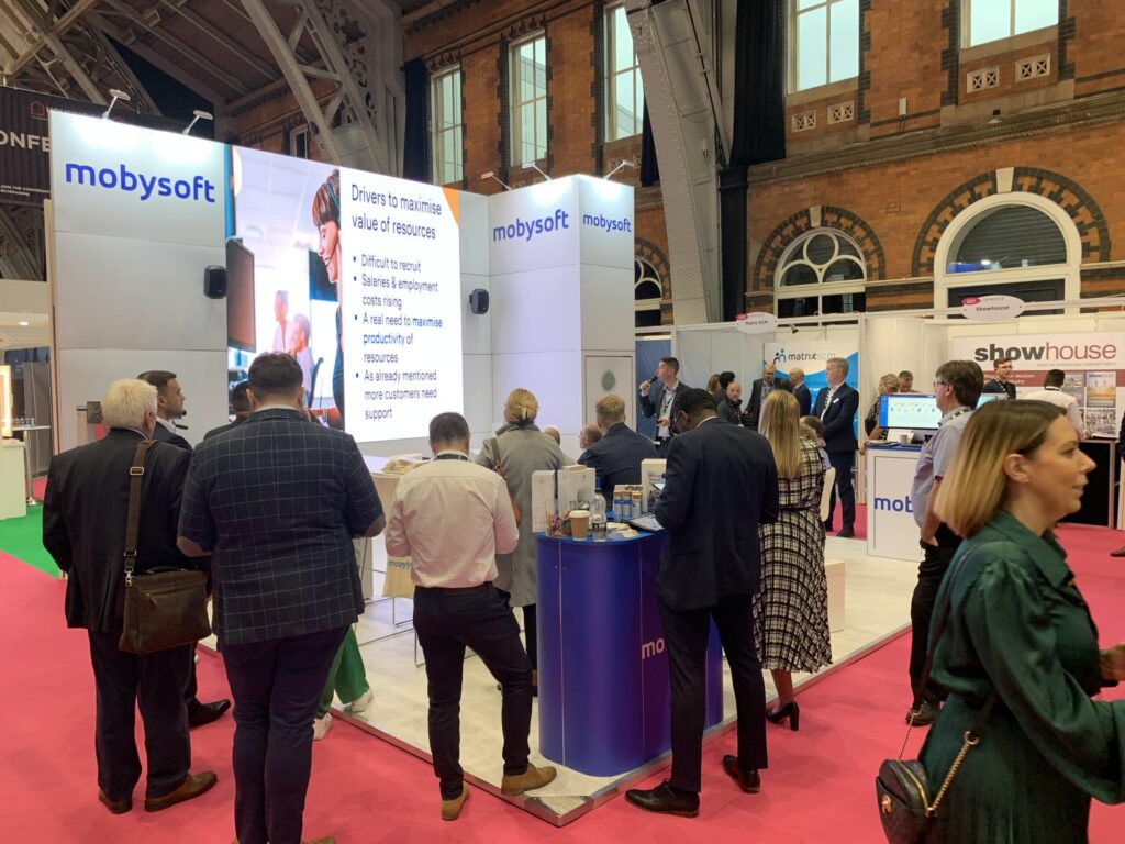 At HOMES 2022, people gather around the Mobysoft booth at a bustling trade show. The booth showcases a screen with key insights, while attendees engage in lively discussions and view displays. In the background, other booths and conference signage add to the vibrant atmosphere. ©Mobysoft