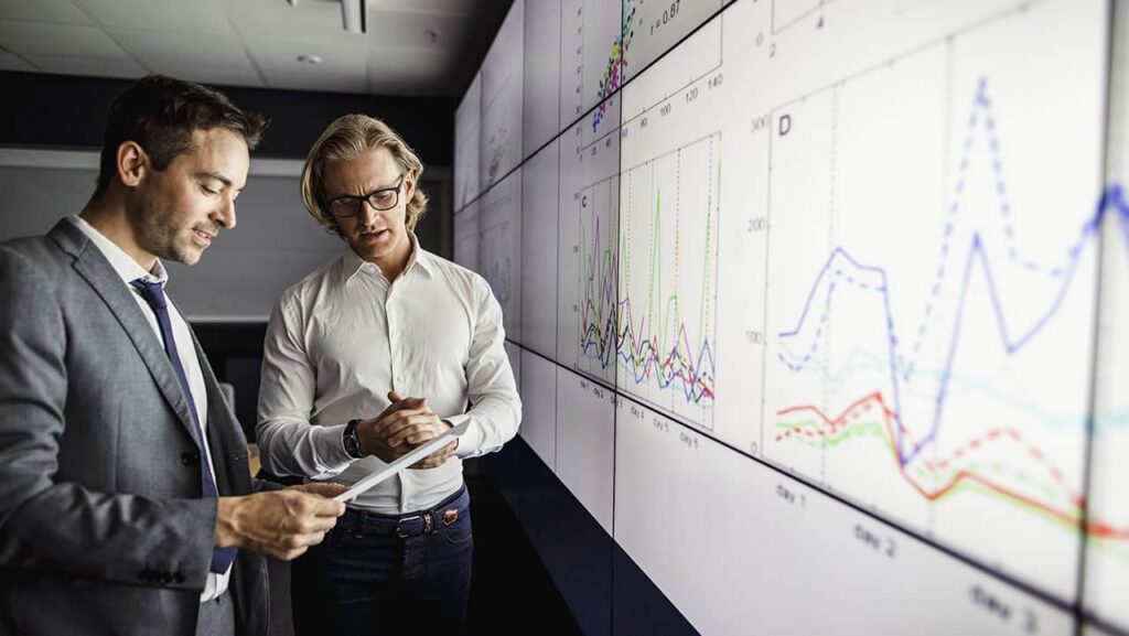 Two men in business attire discuss data displayed on a large screen filled with graphs and charts. One holds a tablet, while both focus on the information, following the golden thread of insight that weaves through their professional analysis and decision-making process. ©Mobysoft