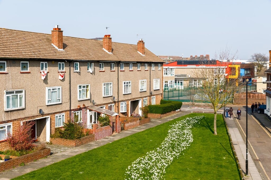A row of brick houses with a manicured lawn and a path lined with white flowers. In the background, there's a modern building and a fenced tennis court. The sky is clear and blue. ©Mobysoft