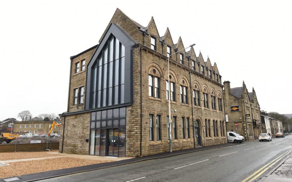 A large, historic stone building with multiple gables and arched windows stands on a street. It features modern glass paneling on one section, highlighting the evolution of Together Housing. A white van is parked nearby, and construction equipment is visible in the background. ©Mobysoft