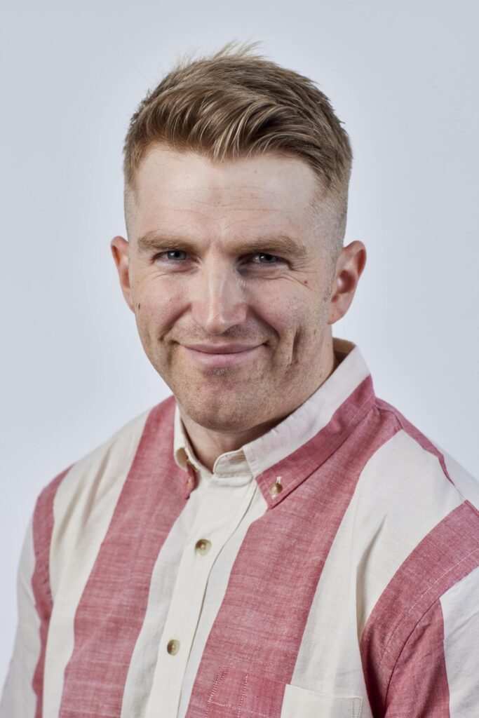 A person with short, light brown hair wearing a red and white striped shirt smiles slightly, standing against a plain gray background that resembles the minimalist design often seen in modern social housing developments. ©Mobysoft