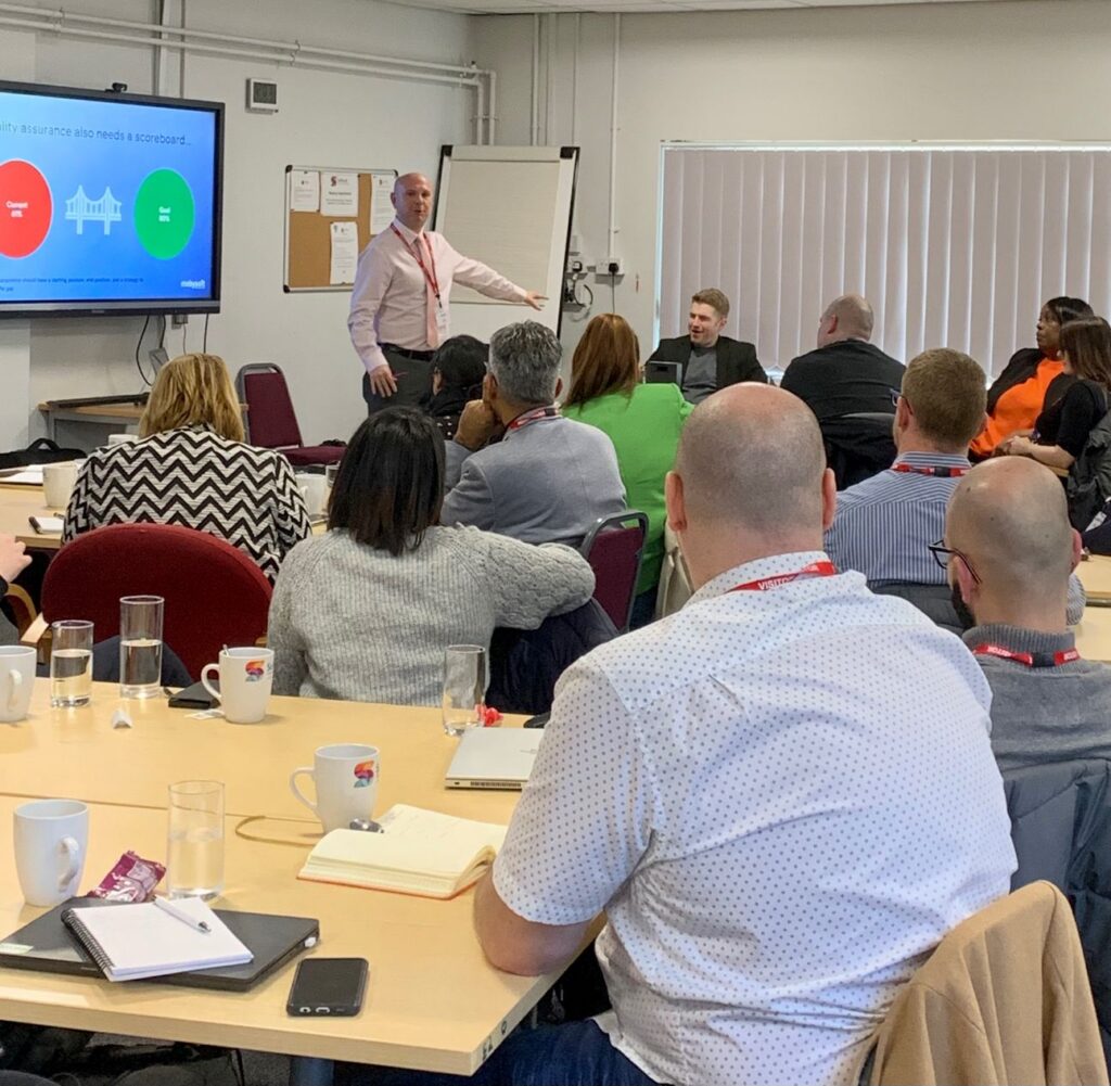 A presenter stands by a screen displaying a presentation slide on social housing in a conference room. Attendees are seated around tables, listening attentively, with notebooks and beverages in front of them. ©Mobysoft