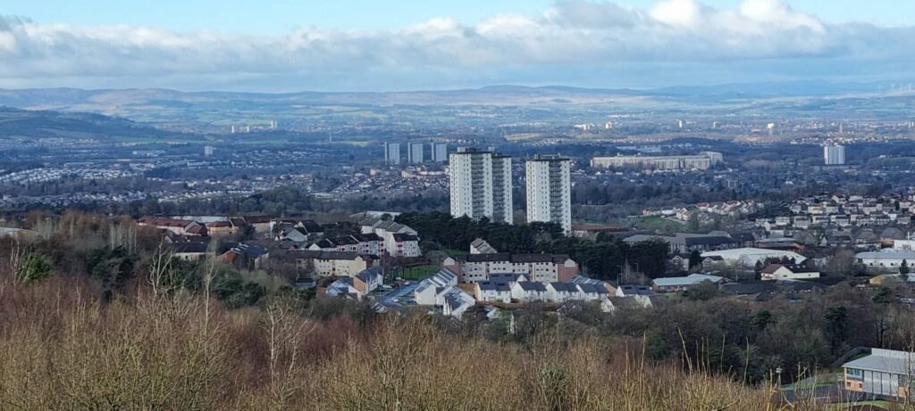 A scenic view of the city showcases a cluster of tall apartment buildings, akin to Cassiltoun Housing, at the center, surrounded by smaller houses and lush trees. In the background, rolling hills stretch under a partly cloudy sky. ©Mobysoft