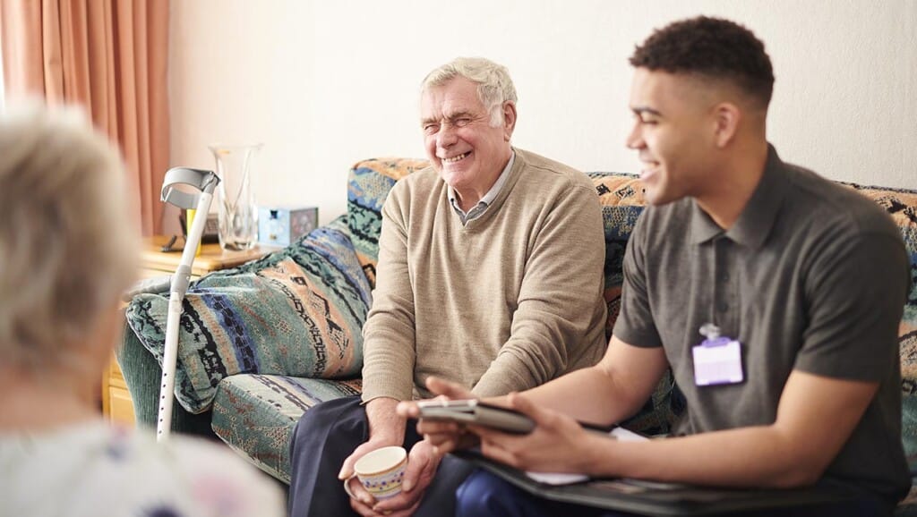 An elderly man smiles while sitting on a couch with a cup in his hand, next to a young man holding a tablet and wearing a name badge. A cane rests against the couch, and a woman sits in the foreground. The setting appears to be a comfortable living room. ©Mobysoft