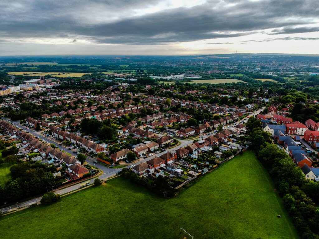 Aerial view of a suburban area with rows of houses surrounded by green fields and trees. The overcast sky looms above the blend of urban and rural landscapes, as Oxford City Council works to address housing needs amidst a backdrop extending into the horizon. ©Mobysoft