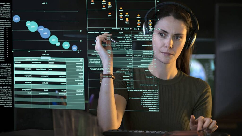 A woman wearing headphones interacts with a transparent digital interface displaying charts and data, analyzing damp trends. She appears focused, with one hand touching the screen and the other on a keyboard. The background is dimly lit. ©Mobysoft