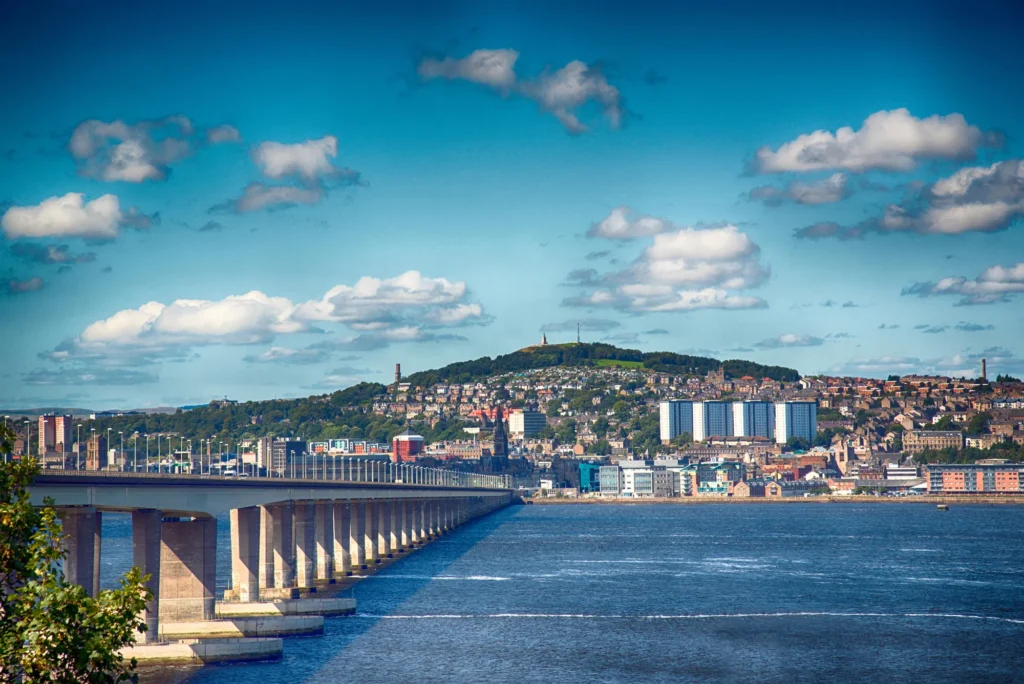 A wide river with a long bridge stretches towards a city on the opposite bank, set against a backdrop of rolling hills. Beneath the scattered clouds in the blue sky, Dundee City Council proudly announces it has reduced arrears by £230K. ©Mobysoft