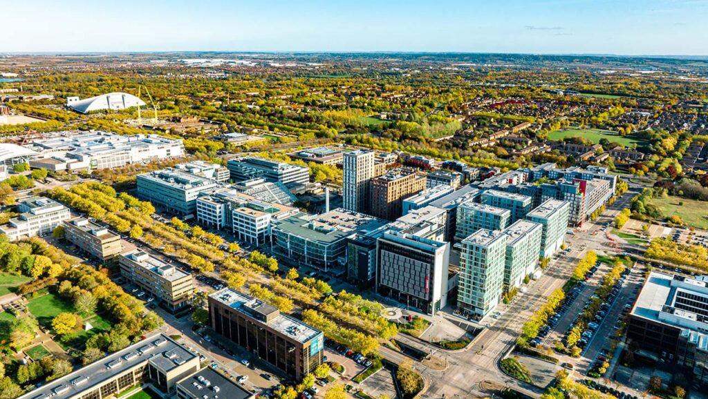 Aerial view of a modern cityscape under Milton Keynes Council, featuring large office buildings surrounded by trees and roads. The landscape extends into the background with residential areas, fields, and a visible sports stadium. The sky is clear and blue. ©Mobysoft