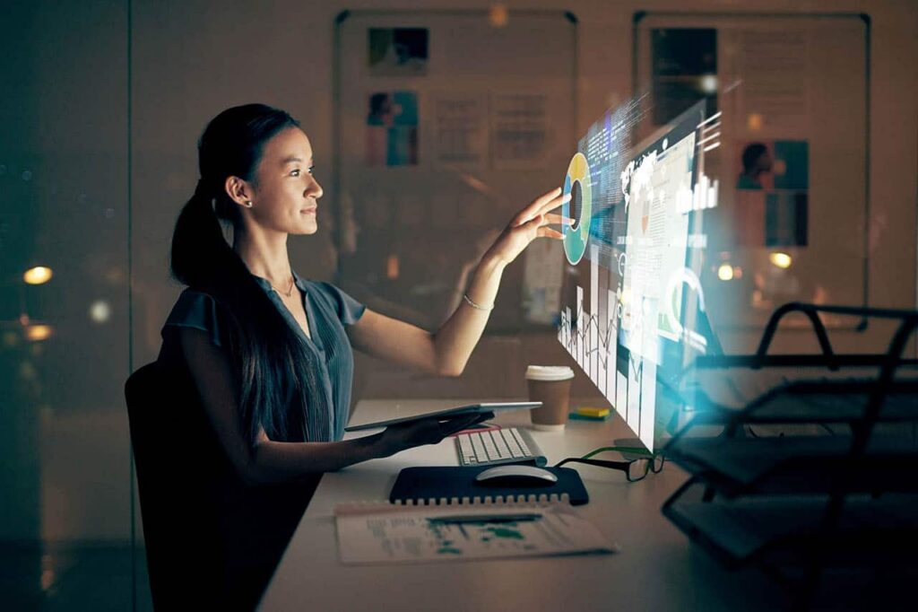 A woman sits at a desk in a dimly lit office, interacting with a touchscreen display like many IT directors do. The screen shows charts and data visualizations. A notebook, pen, and a cup are on the desk. Shelves with files are in the background. ©Mobysoft