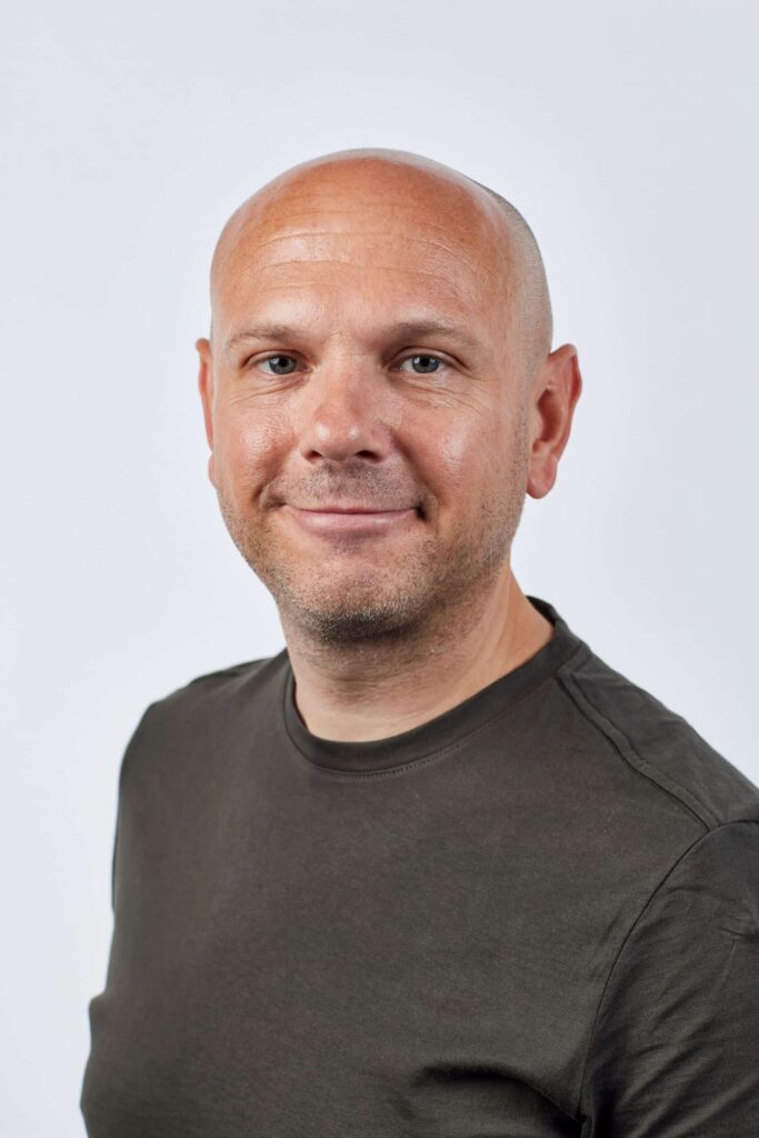 A person with a shaved head wearing a dark green t-shirt, reminiscent of the serene tones found in social housing designs, smiles slightly against a plain white background. ©Mobysoft