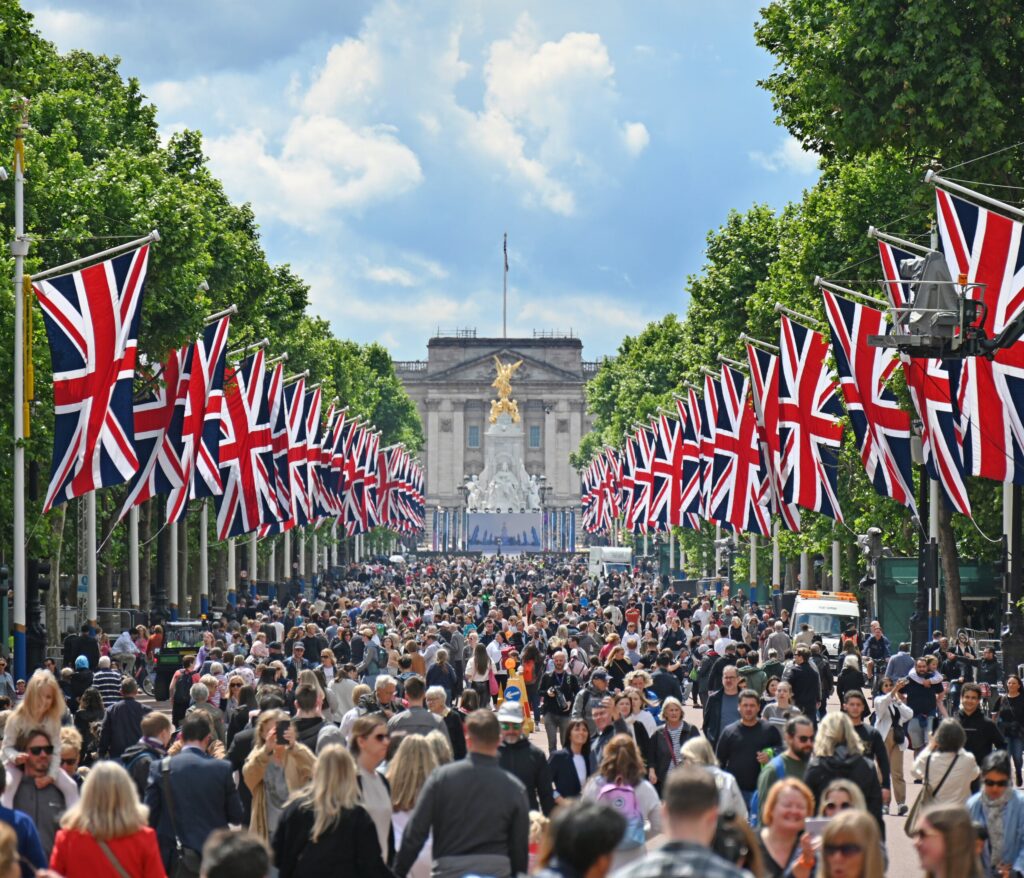A large crowd fills a tree-lined avenue adorned with numerous Union Jack flags, celebrating the coronation. In the background, a grand building with an ornate golden statue on top is visible under a partly cloudy sky. ©Mobysoft