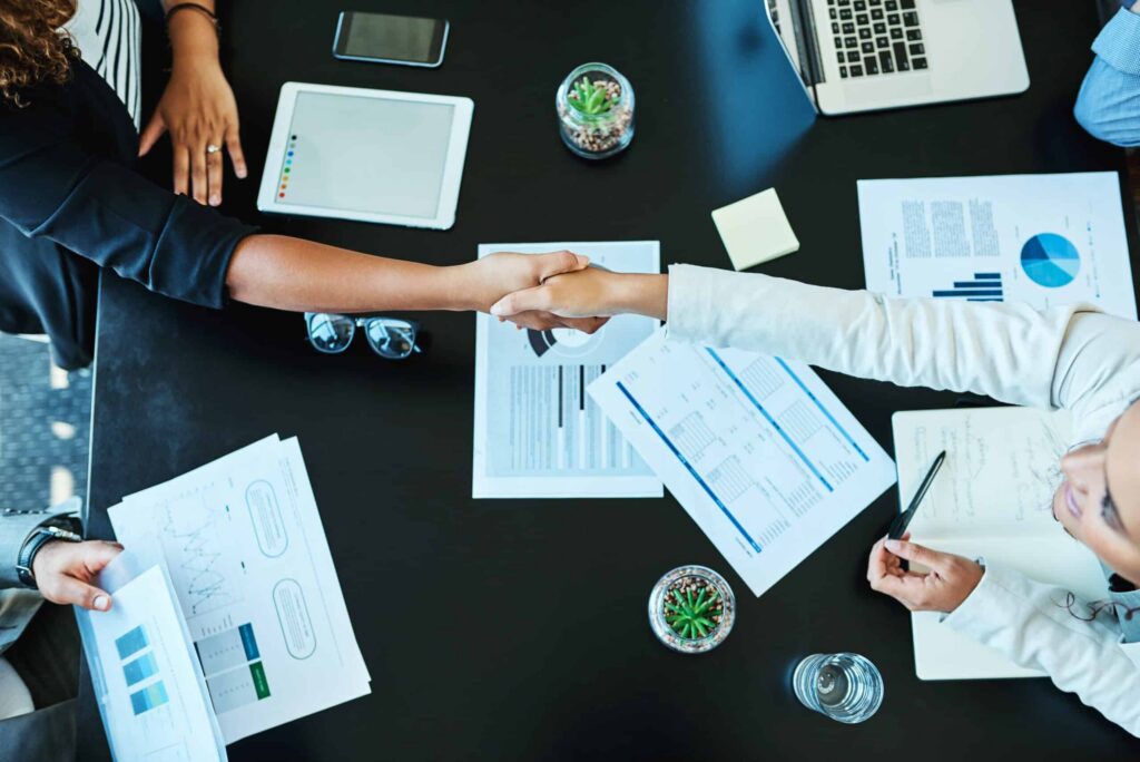Two people shaking hands over a black table with business documents, a tablet, and a laptop signifies the finalization of mergers within social housing. Notebooks, charts, a small plant, and a glass of water complete this professional setting. ©Mobysoft