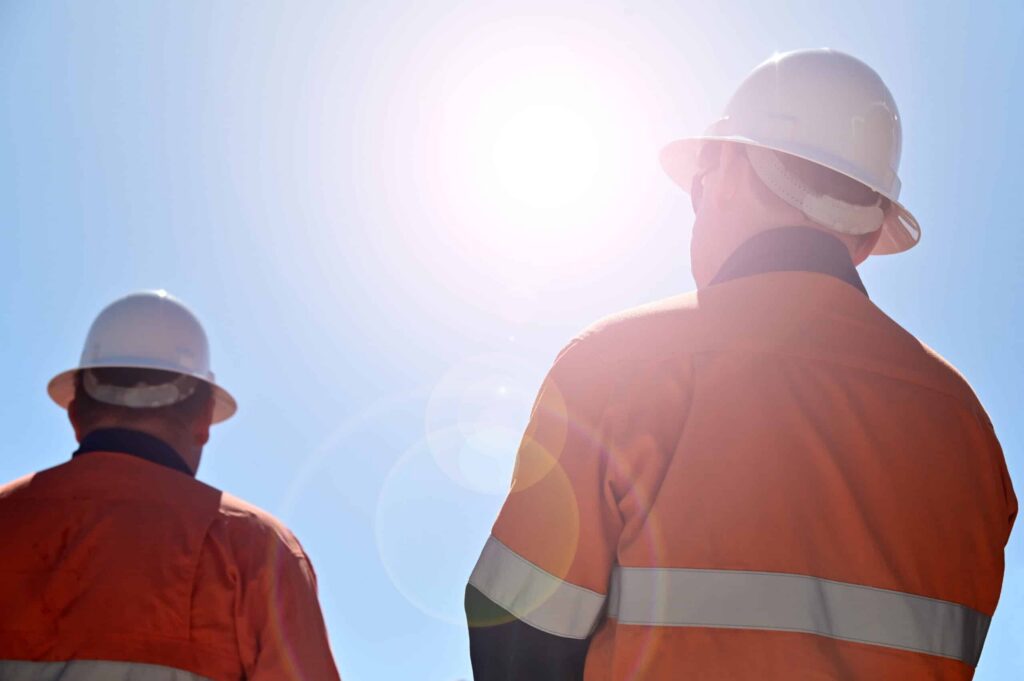Two construction workers in orange safety jackets and white helmets look towards the bright sun in a clear blue sky, lens flare visible, while delivering repairs and maintenance services during summer. ©Mobysoft