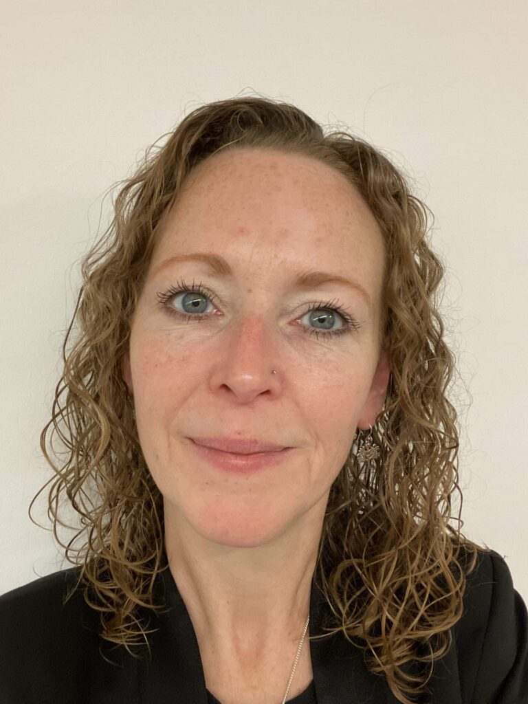 A person with curly, shoulder-length hair and blue eyes, wearing a black top, looks into the camera with a neutral expression. The setting is simple and direct, like the straightforward design often seen in social housing projects. ©Mobysoft