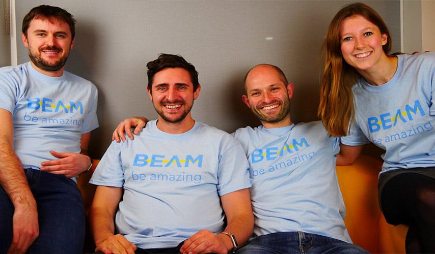Four people wearing light blue t-shirts with "BEAM be amazing" printed on them are sitting on a couch, smiling at the camera. The background is a plain wall, and they appear to be in a casual setting. ©Mobysoft