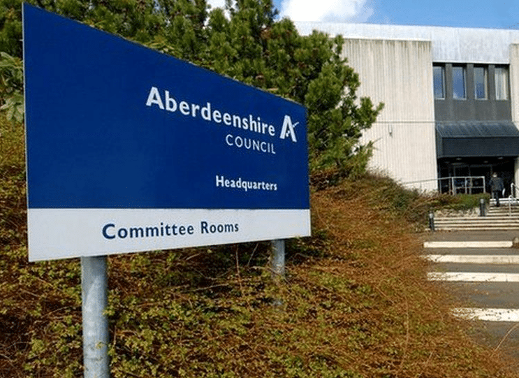 A sign reads "Aberdeenshire Council Headquarters, Committee Rooms" in front of a sleek, modern Aberdeenshire building surrounded by large steps and lush greenery. ©Mobysoft