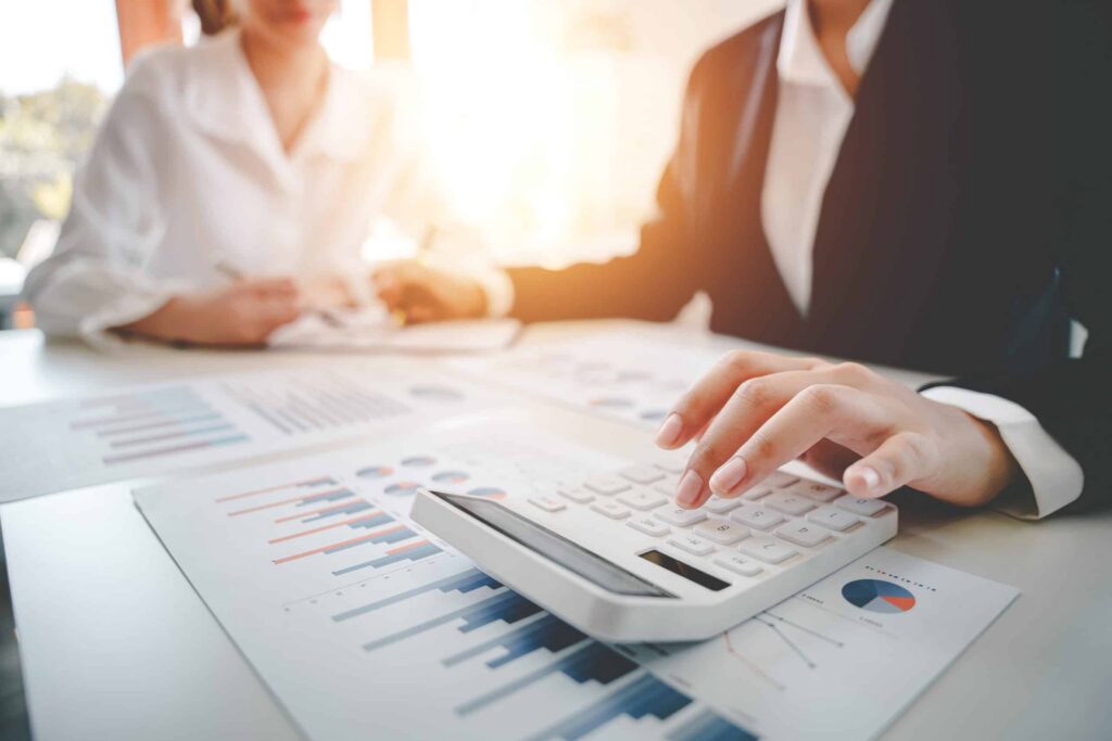 Two business professionals analyze the financial viability of housing associations as they review reports. One uses a calculator, while charts and graphs are spread across the table. The office setting is bathed in warm, natural light, fostering a focused yet relaxed atmosphere. ©Mobysoft