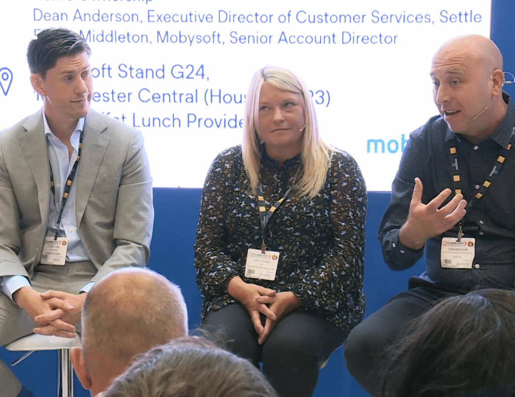 Three individuals are seated and engaged in conversation at a panel discussion. A screen behind them displays information about a partnership event. They wear conference badges, indicating participation in a professional event. ©Mobysoft