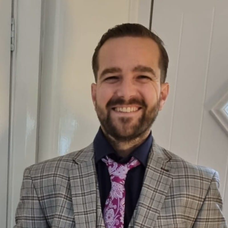 A man with a beard smiling, wearing a checkered suit jacket, dark shirt, and patterned pink tie, stands indoors in front of a white door. ©Mobysoft