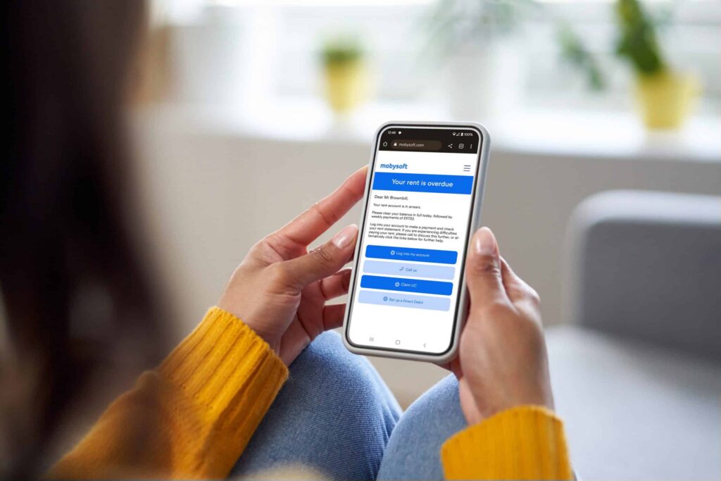 A person wearing a yellow sweater is holding a smartphone displaying a webpage with a message about overdue rent. The screen shows options for payment and assistance. The scene is indoors with a bright window in the background. ©Mobysoft