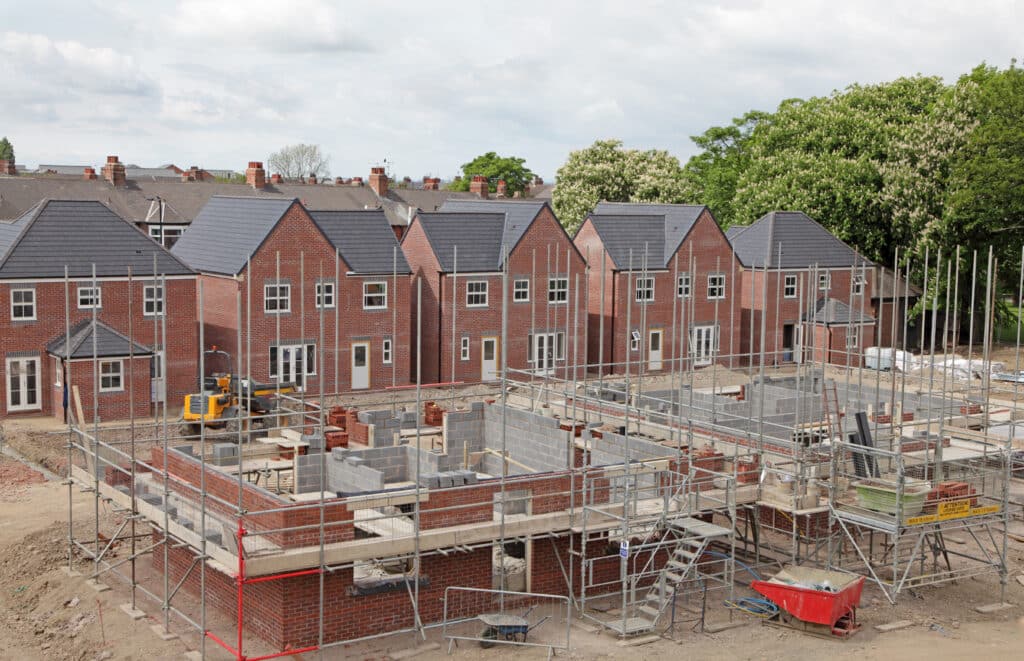 A row of completed, identical brick houses with dark roofs stands behind a busy construction site buzzing with activity. The partially built structures and scaffolding hint at new social housing developments. Trees and a cloudy sky form the backdrop to these unfolding news stories. ©Mobysoft