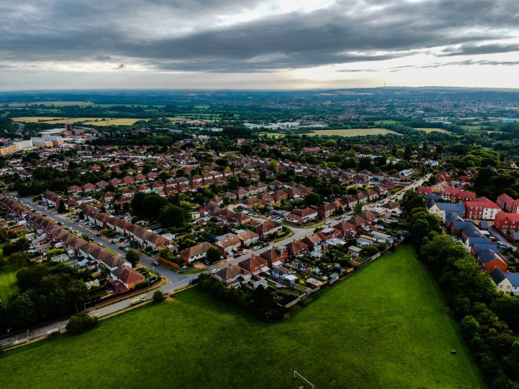 Aerial view of a National Housing Federation residential area with rows of houses and green spaces. The landscape includes trees, fields, and distant hills under a cloudy sky, crafting a serene suburban scene. ©Mobysoft