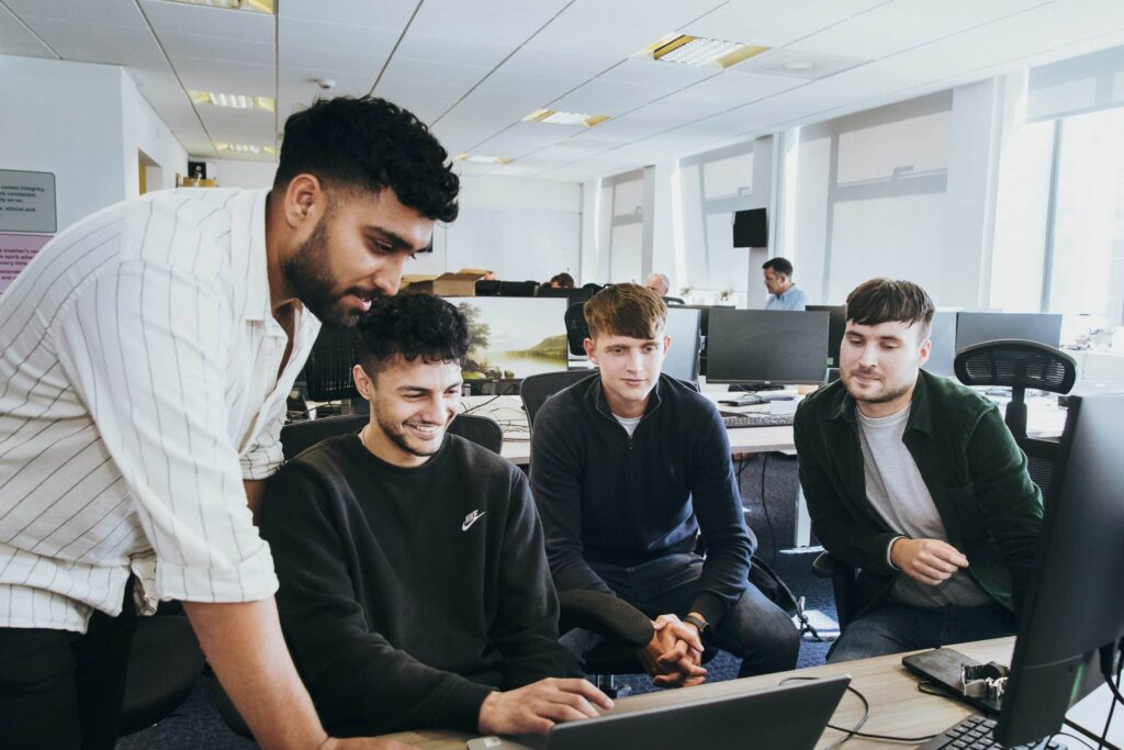 A group of young men gathered around a computer in an office setting. One man stands pointing at the screen while the others, demonstrating staff buy-in for AI, are seated and attentively focused on the display. The office is filled with desks and computer monitors. ©Mobysoft