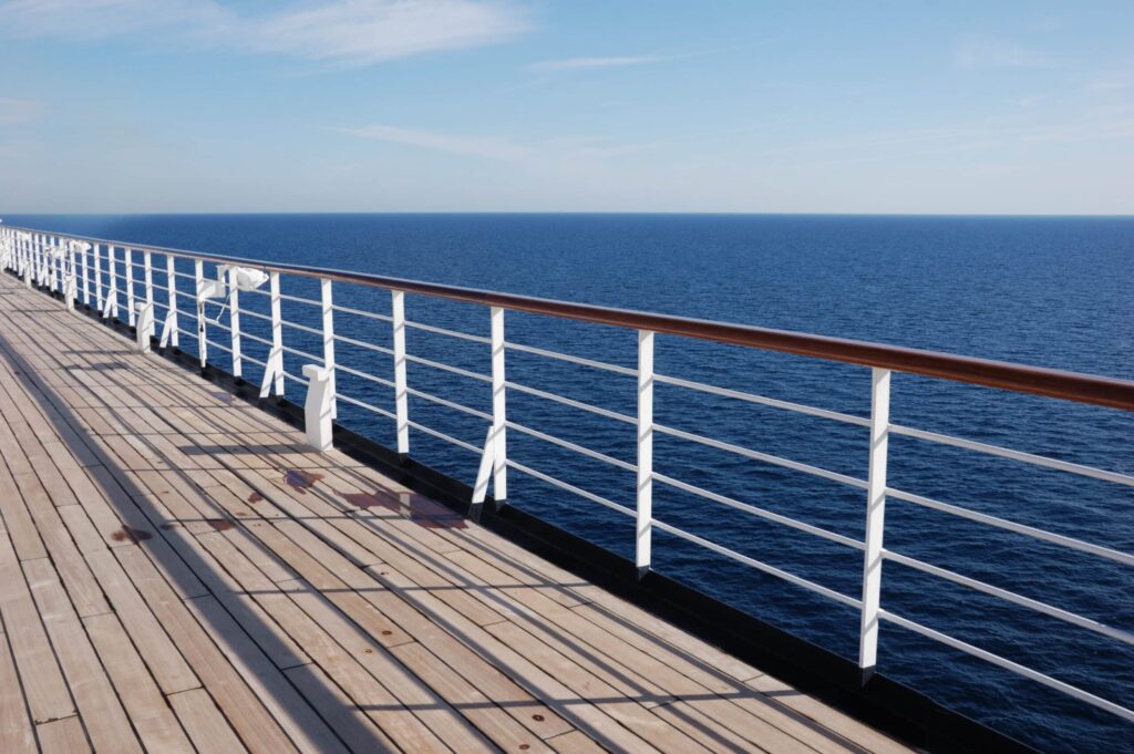 A wooden deck on a cruise ship stretches alongside a white railing, overlooking a vast, calm blue ocean under a clear sky. The sun casts shadows, reminiscent of AI guardrails, across the deck. ©Mobysoft