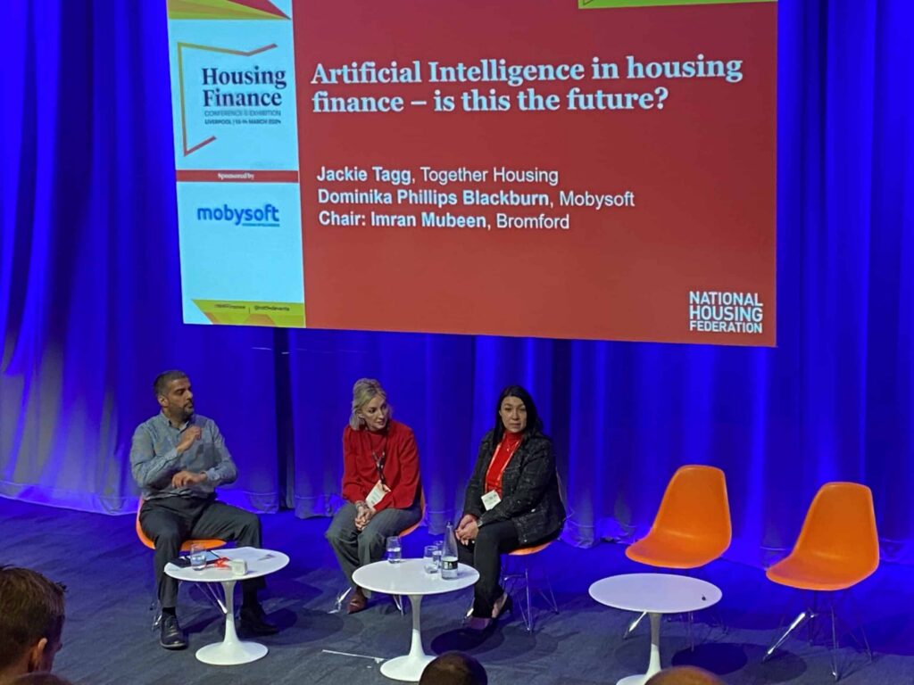 Three panelists seated on stage at a conference discuss "Artificial Intelligence in Housing Finance – is this the future?" The screen flashes the topic and participant names, while a blue curtain backdrop frames the scene with empty orange chairs awaiting their insights. ©Mobysoft