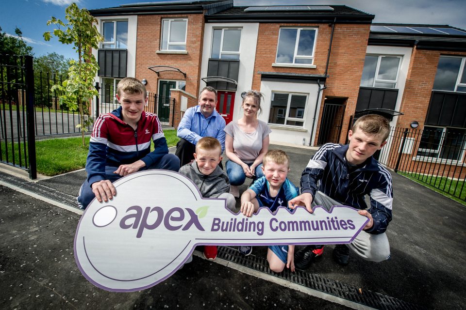 A family of six beams with joy in front of their new home in Northern Ireland, clutching a large key-shaped sign that reads "Apex Building Communities." They stand on a paved surface, with the charming house and garden visible behind them. ©Mobysoft
