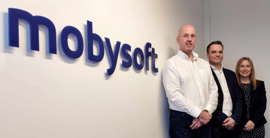 Three people stand smiling in front of a wall adorned with the "Mobysoft" logo. The Executive Management Team includes two men and one woman, all dressed in professional attire. ©Mobysoft