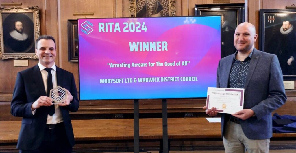 Two men stand in front of a screen displaying "RITA 2024 Winner: Arresting Arrears for the Good of All". One holds a trophy, the other a certificate. They are in a wood-paneled room with historical portraits on the walls. ©Mobysoft
