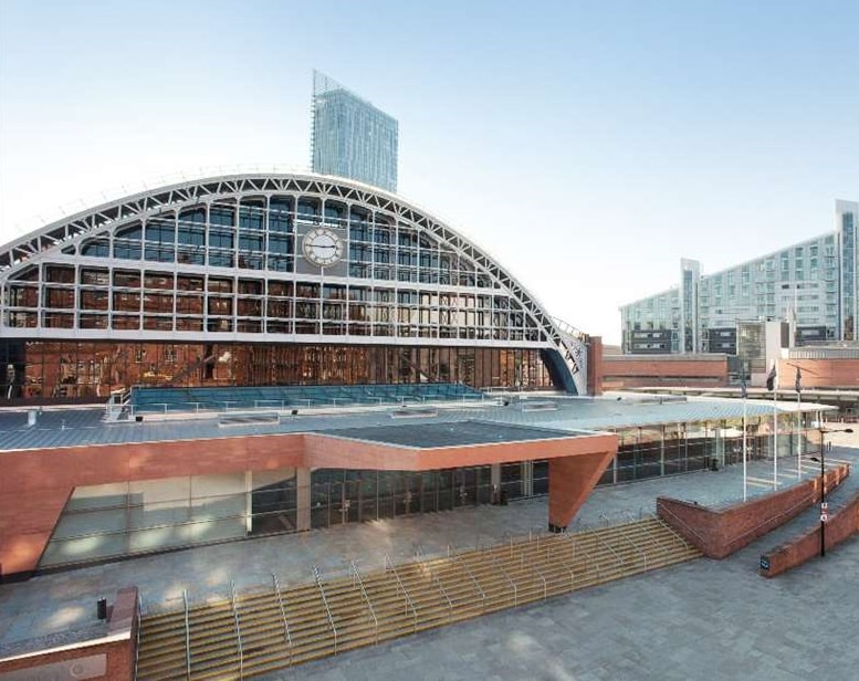 Modern architectural building with a large arched glass facade and a clock at the top. The structure, part of the innovative Housing 2024 development, features red brick and glass elements with steps leading to the entrance. A tall skyscraper is visible in the background against a clear sky. ©Mobysoft