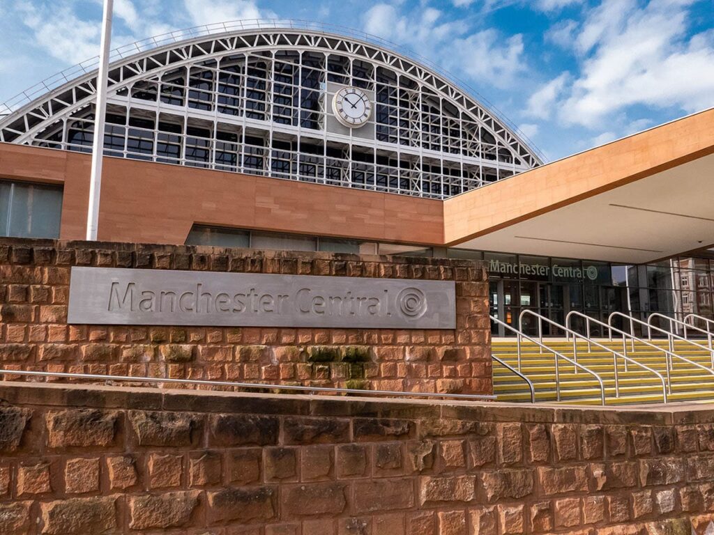 Exterior view of Manchester Central Convention Complex. The building features a large arched roof with a clock at the center. A sign reads "Manchester Central." Steps lead up to the entrance. The sky is partly cloudy. ©Mobysoft