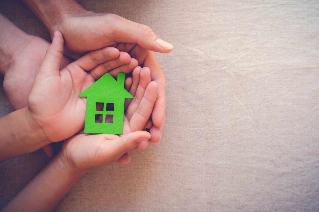 Close-up of adult and child hands holding a small green house cutout together on a beige background, representing home and family support. ©Mobysoft