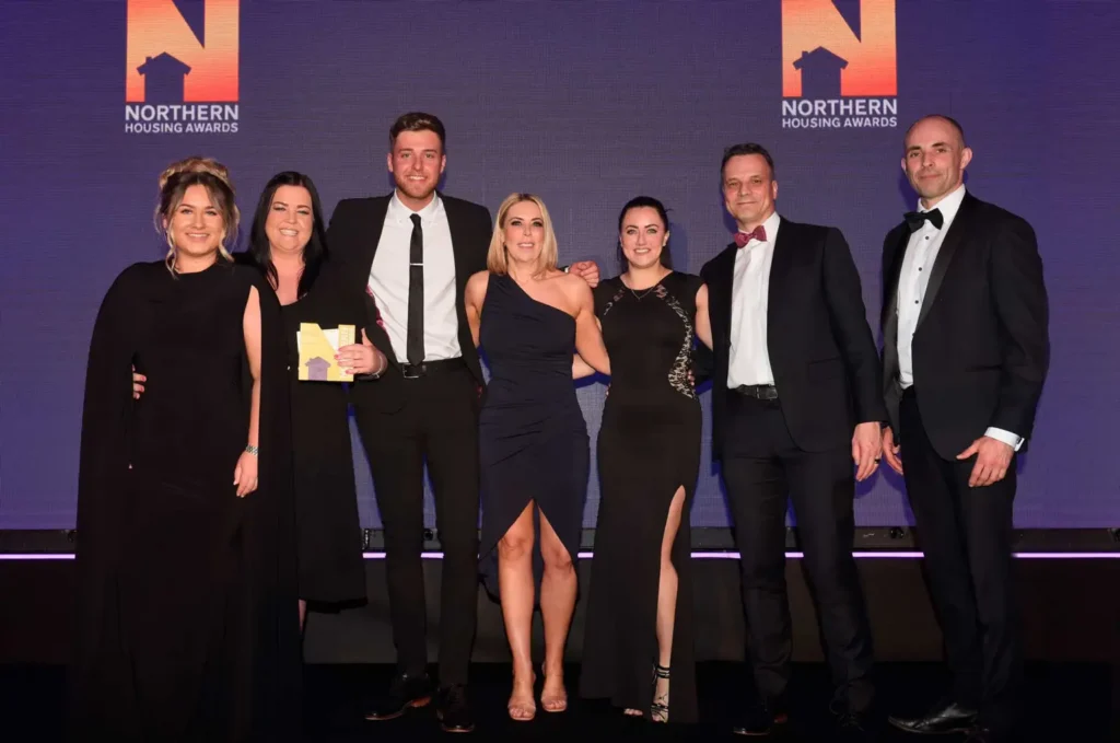 A group of seven people from Karbon Homes, dressed formally, pose for a photo at the Northern Housing Awards. Three women and four men stand shoulder to shoulder, some proudly holding awards. The background prominently displays the event's logo. ©Mobysoft