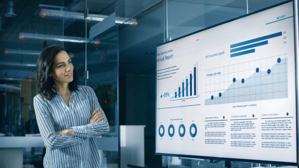 A person stands in an office setting, closely examining a large screen displaying a detailed financial report with charts and graphs. The comprehensive data strategy is evident through the line graph, bar graphs, and circular diagrams, keeping the individual engaged and thoughtful. ©Mobysoft
