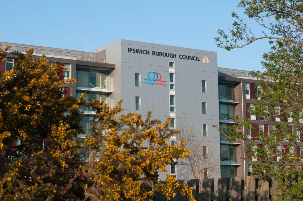 A modern building with the "Ipswich Borough Council" sign is partially obscured by lush trees. The facade proudly bears a "smarter working" logo, under a clear blue sky. ©Mobysoft