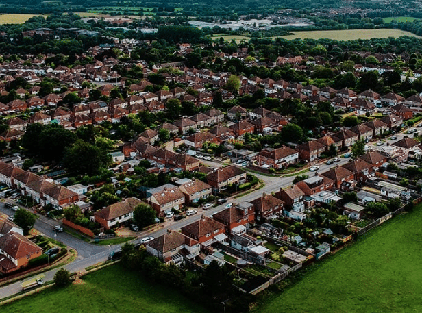 From an aerial view, the residential neighborhood showcases rows of houses seamlessly integrated with lush greenery and roads. The landscape features fields and trees, striking a balance between urban and rural areas, embodying the concept of data-driven social housing repairs for sustainable development. ©Mobysoft