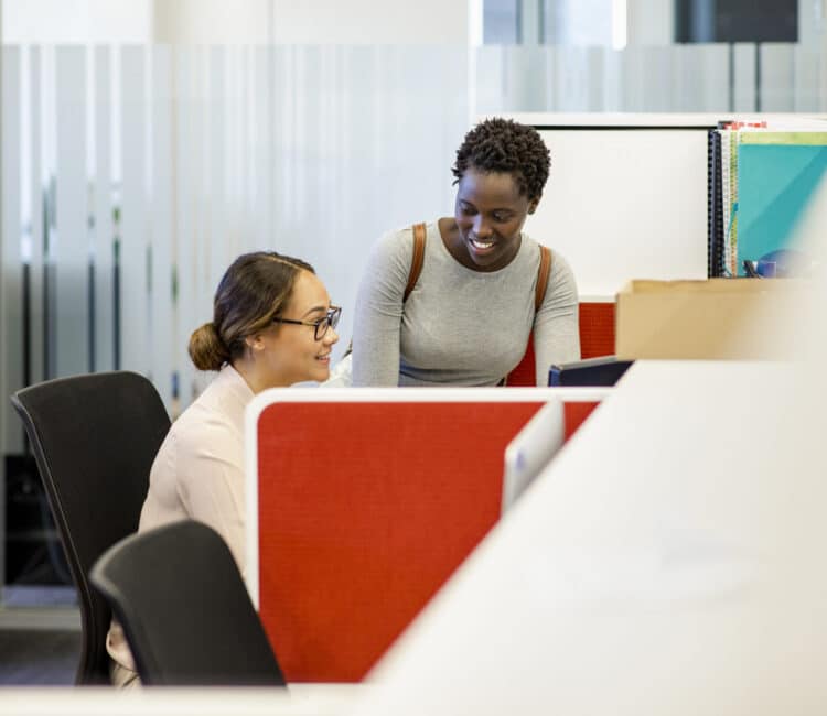 Two colleagues in an office setting, one sitting at a desk with a computer and the other standing nearby, smiling and engaging in conversation. The office features red partitions and glass panels in the background. ©Mobysoft