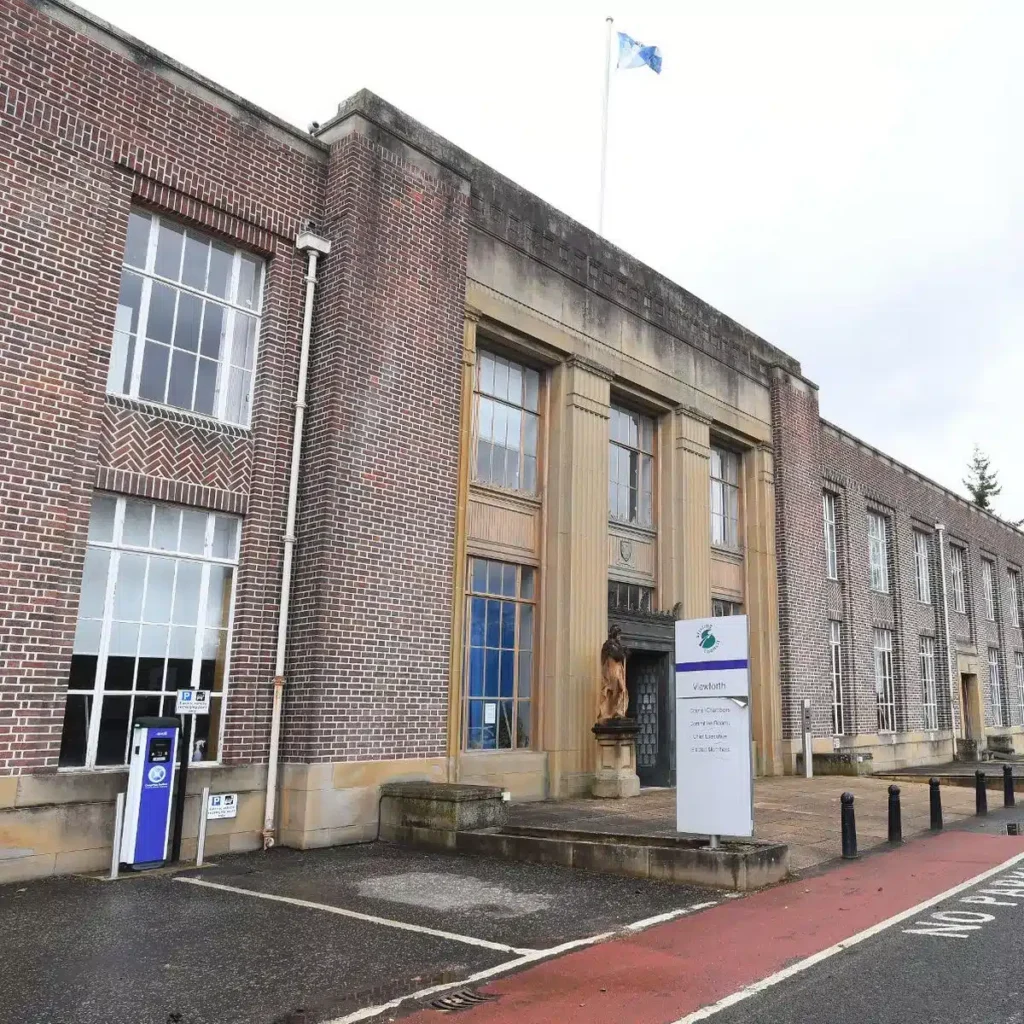 A large brick building with tall windows and a central entrance, featuring a flag on the roof. There's a sign and a statue near the entrance. A bike lane runs in front, and the sky is overcast. ©Mobysoft