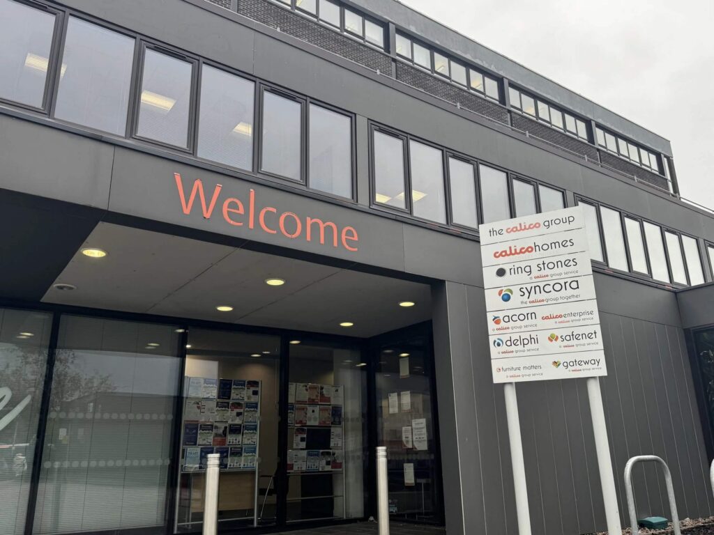 A modern office building with a glass entrance proudly displays "Welcome" in orange above the door. Listings like the Calico Group and South Yorkshire Housing Association adorn a sign. The structure boasts multiple windows and a sleek gray facade, inviting all who pass by. ©Mobysoft