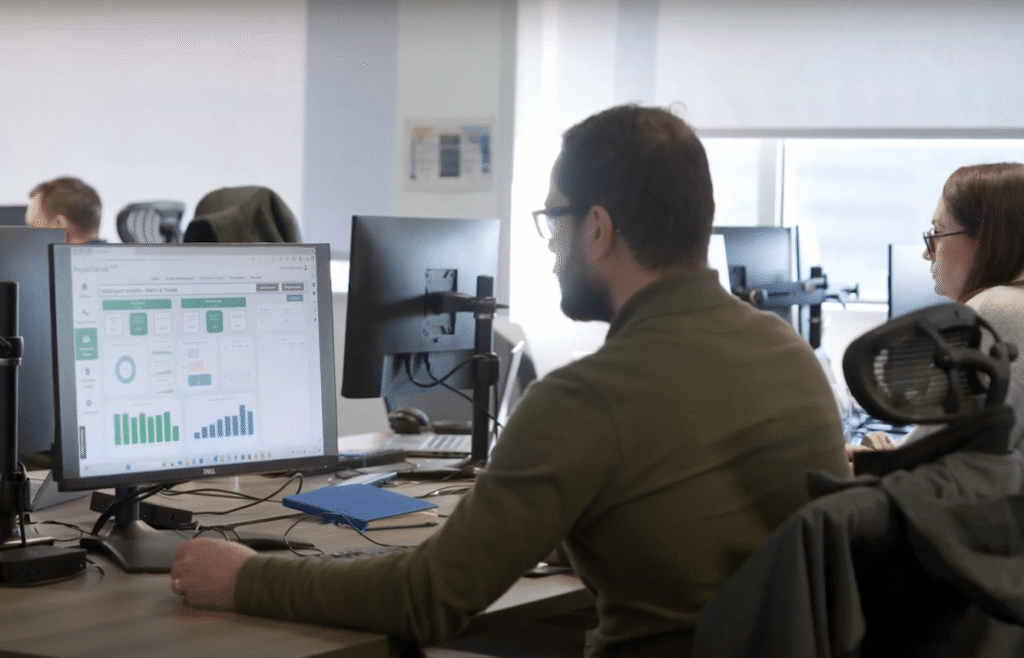 A man in a green shirt is seated at a desk, using repair management software for social housing on his computer displaying graphs and charts. Another person works beside him. The office is filled with multiple desks and chairs equipped with monitors. ©Mobysoft
